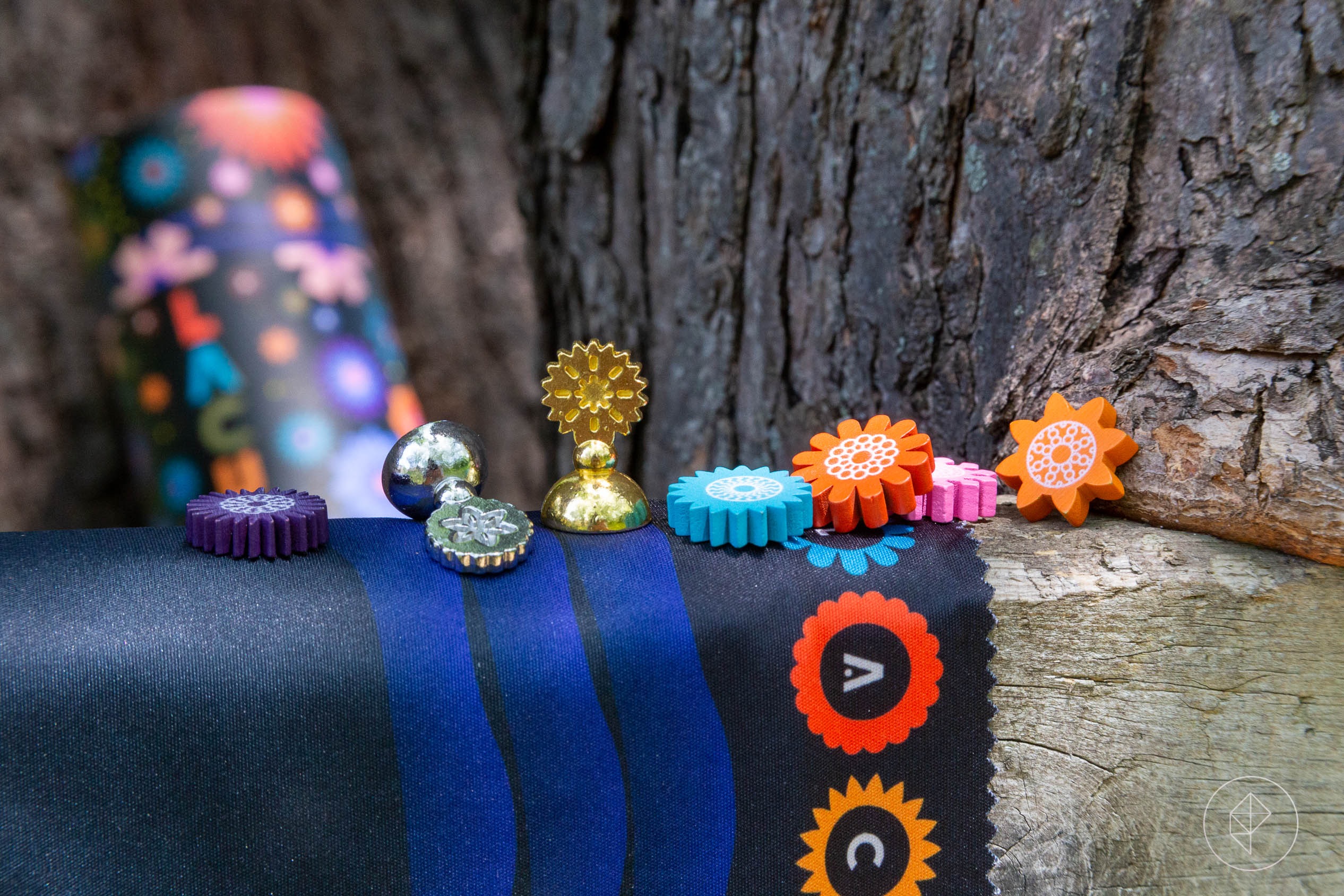 Bits from Lacuna strong across an old treehouse step, the silver maple growing around its edges. The flowers are purple, blue, pink, and orange. The player pawns are gold and silver circles on a small plinth. The tube that is the box for the game is in the background.