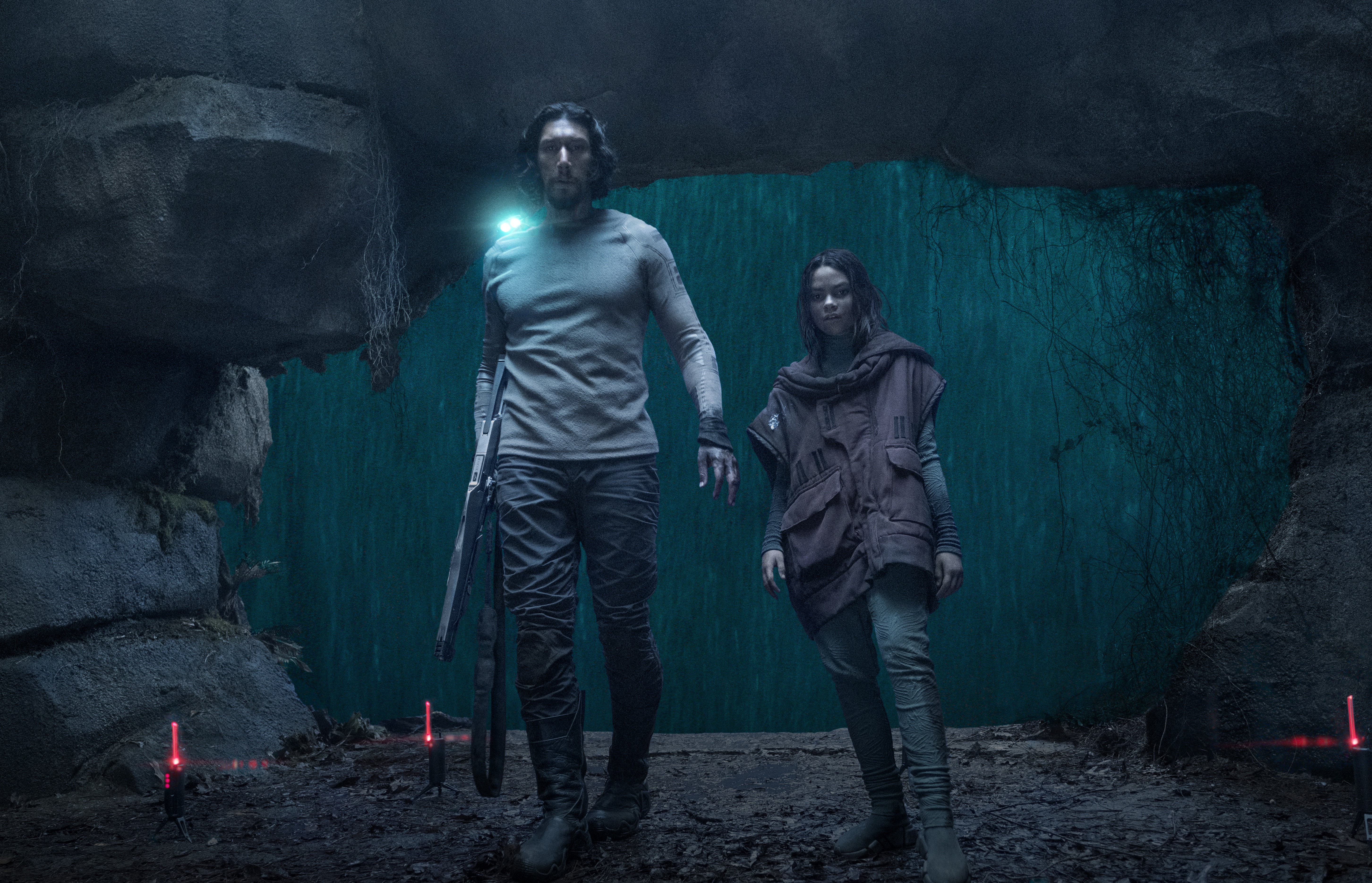 Adam driver’s Mills, wearing a shoulder light and carrying a futuristic rifle, walks with his young brunette teen companion through a cave on a rainy night, setting up a perimeter of red lights in the movie 65