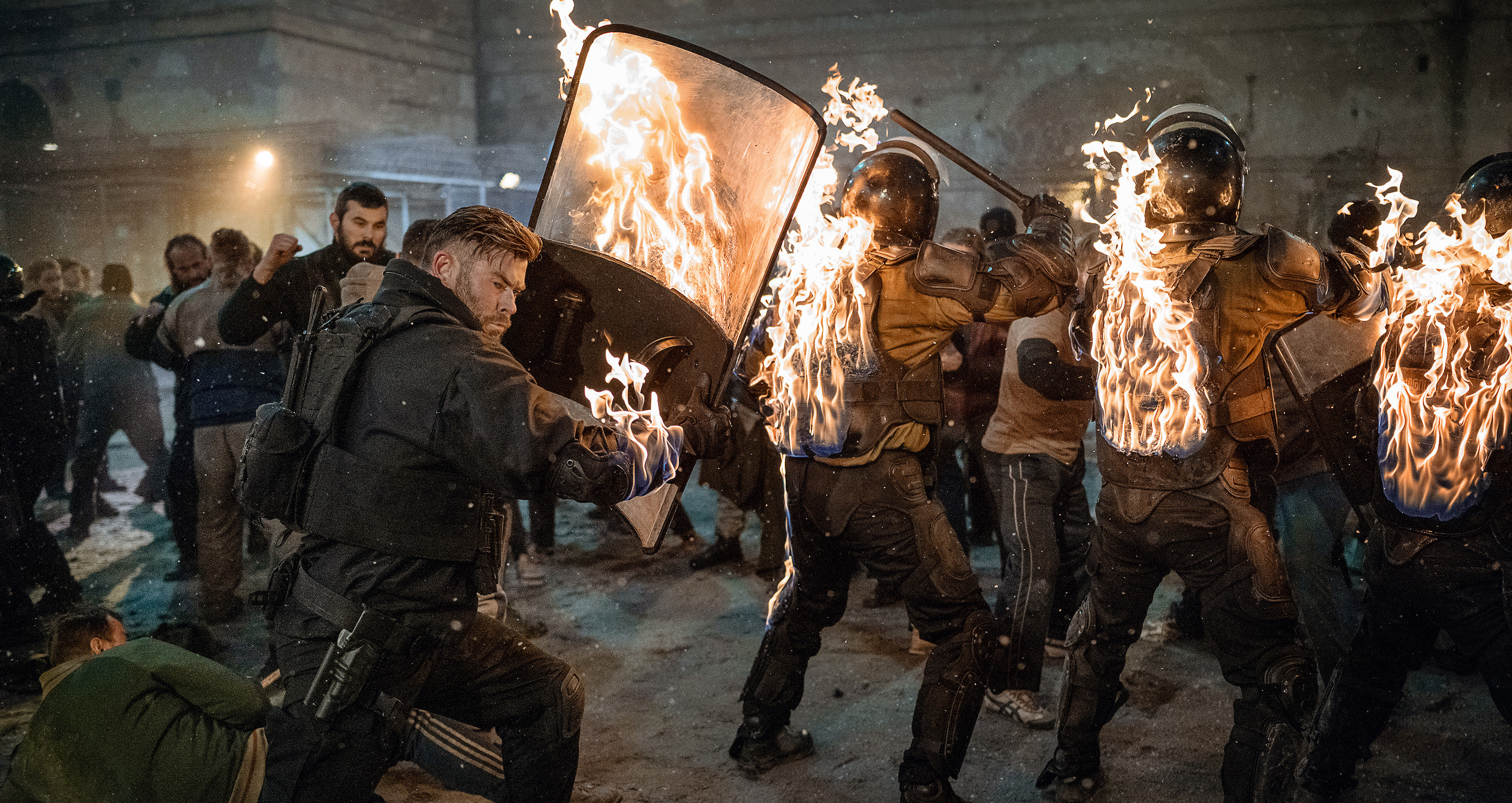 Mercenary Tyler Rake (Chris Hemsworth), with his coat sleeve on fire, charges three armored soldiers holding up riot shields, which are also on fire, in Extraction 2’s astonishing 21-minute handheld action sequence