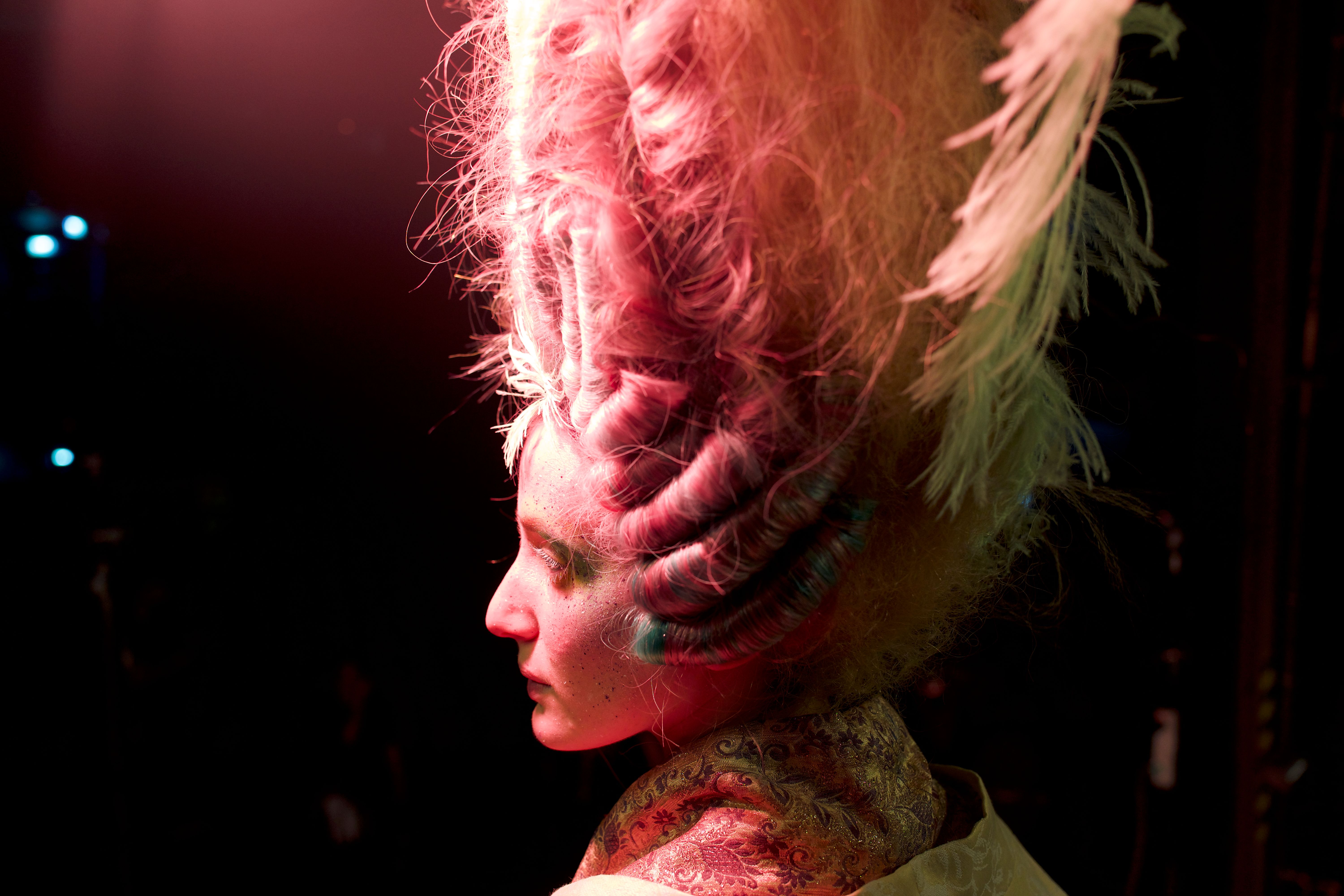 A model in an incredibly elaborate vertical hairdo sits under lights that turn her bright pink and yellow in a shot from A24’s Medusa Deluxe
