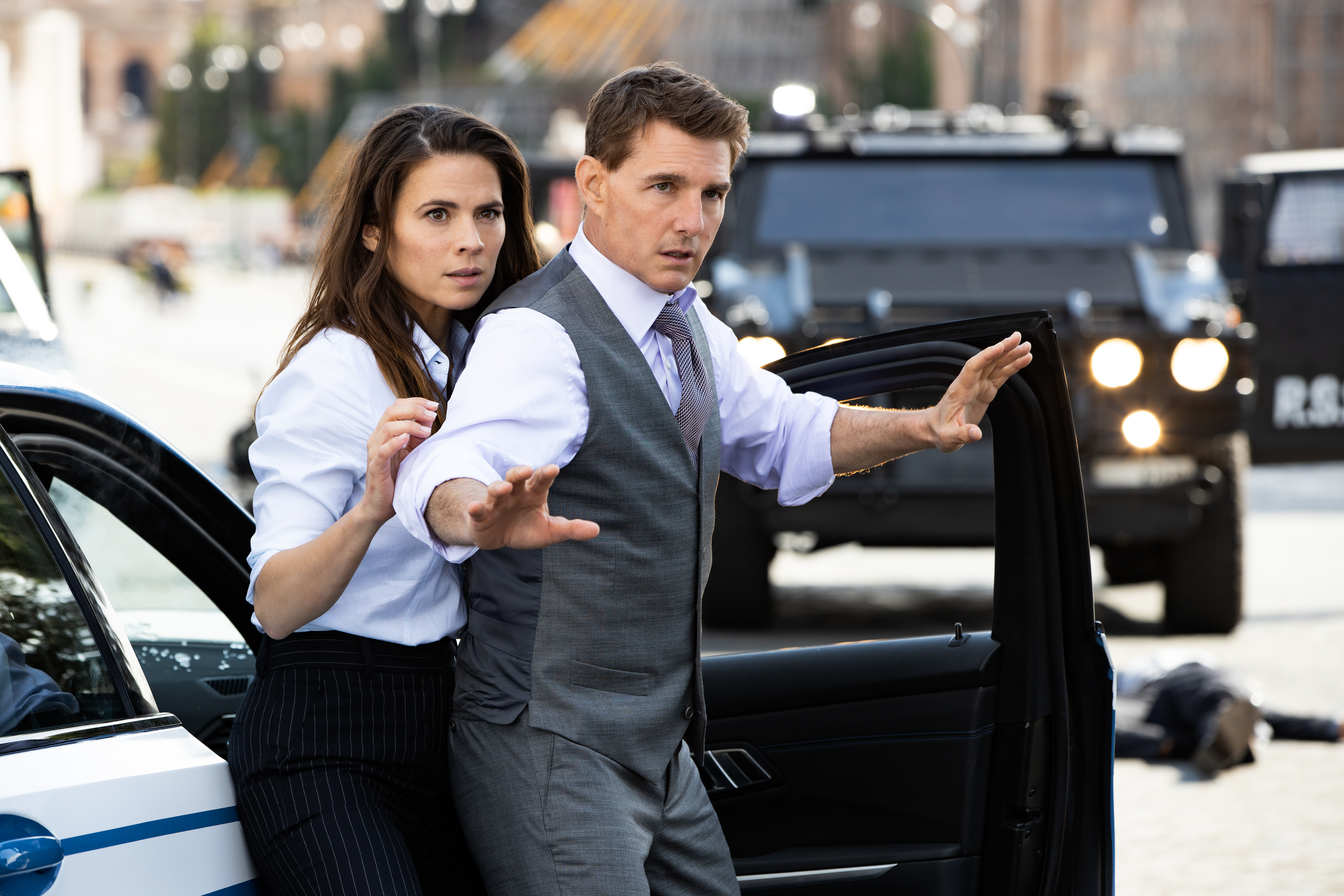 Tom Cruise stands up with his hands raised next to a car with Hayley Atwell standing nervously behind him in a photo from Mission: Impossible - Dead Reckoning Part 1