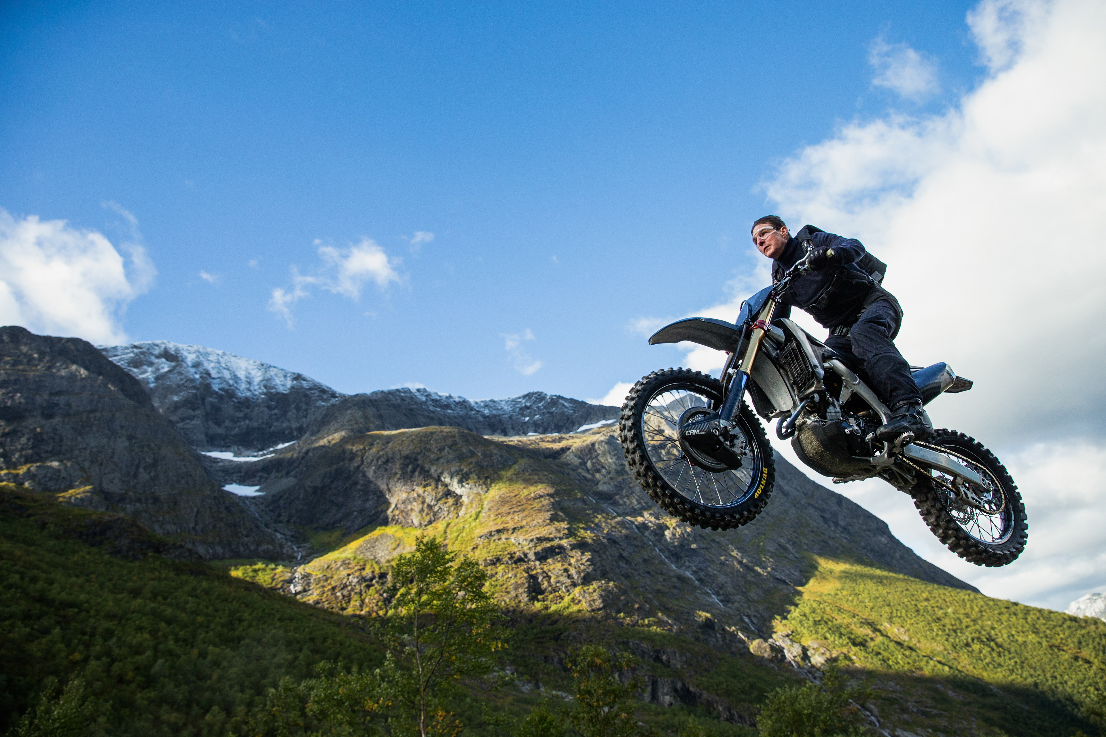 Tom Cruise as Ethan Hunt rides a motorcycle mid-jump off a mountain with the blue sky filling the top half of the screen in a scene from Mission: Impossible - Dead Reckoning Part 1