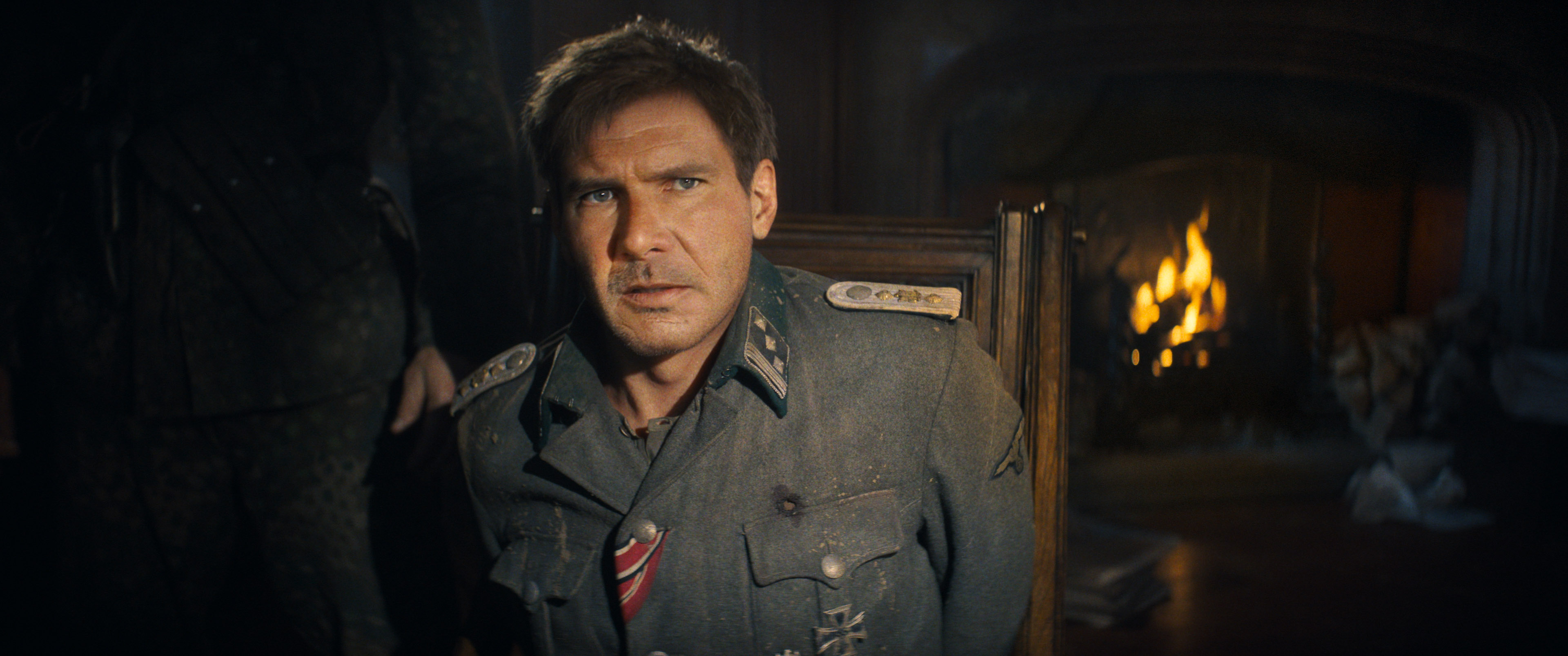 Harrison Ford as Indiana Jones, looking about 40 years old due to CG de-aging, and wearing a grey suit with random American-flag fabric details, looks very concerned about something happening offscreen in Indiana Jones and the Dial of Destiny
