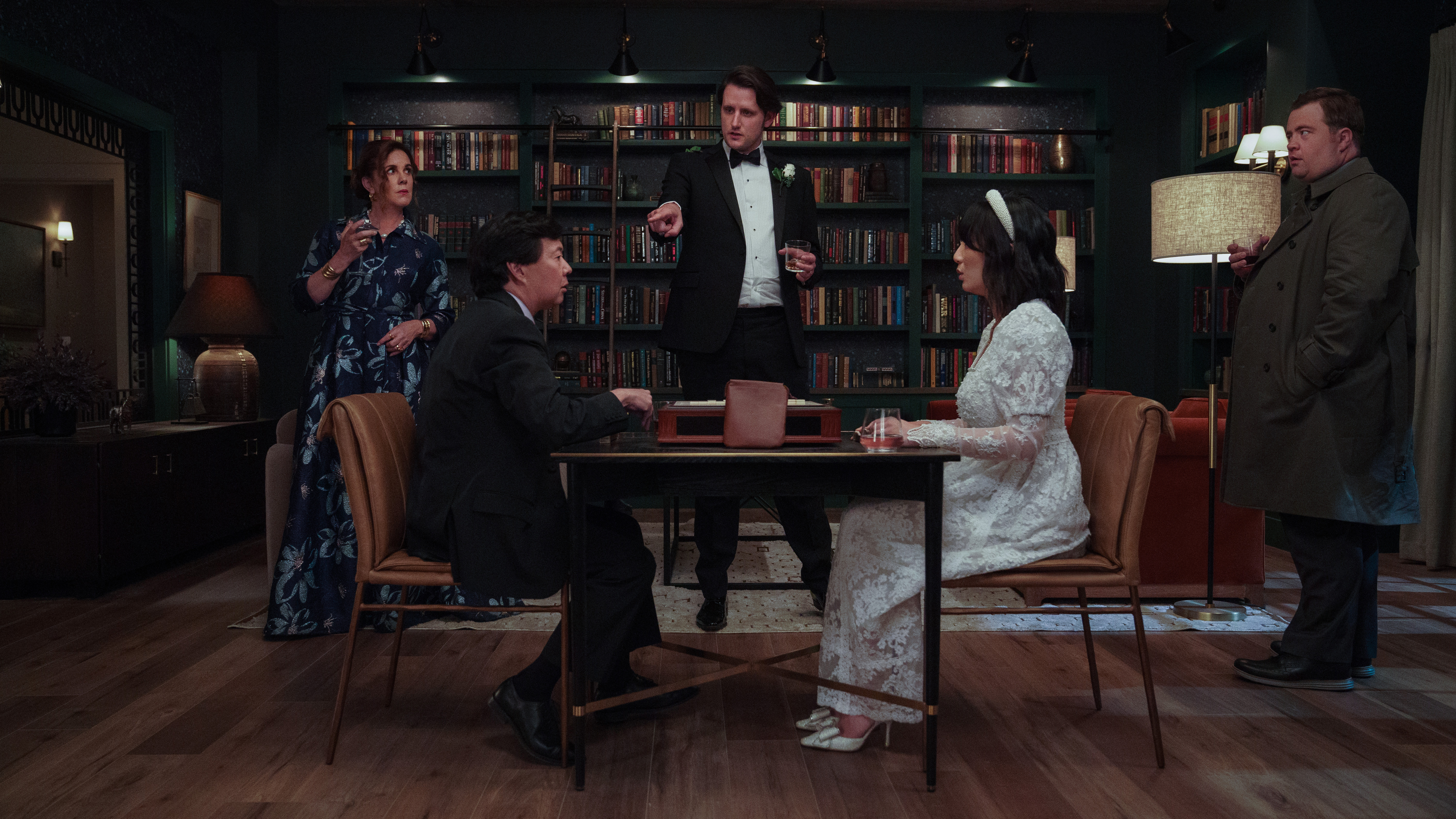 Edgar, a groom in a tux, stands accusing another man seated at a table in front of him across from his bride in a crowded library in season 2 of The Afterparty