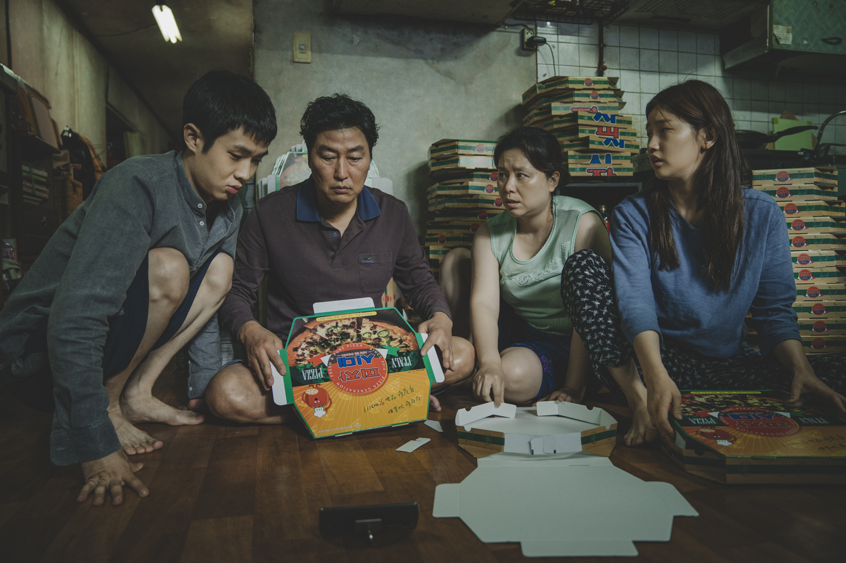 a korean son, dad, mother, and daughter sit in their tiny apartment folding pizza boxes in Parasite