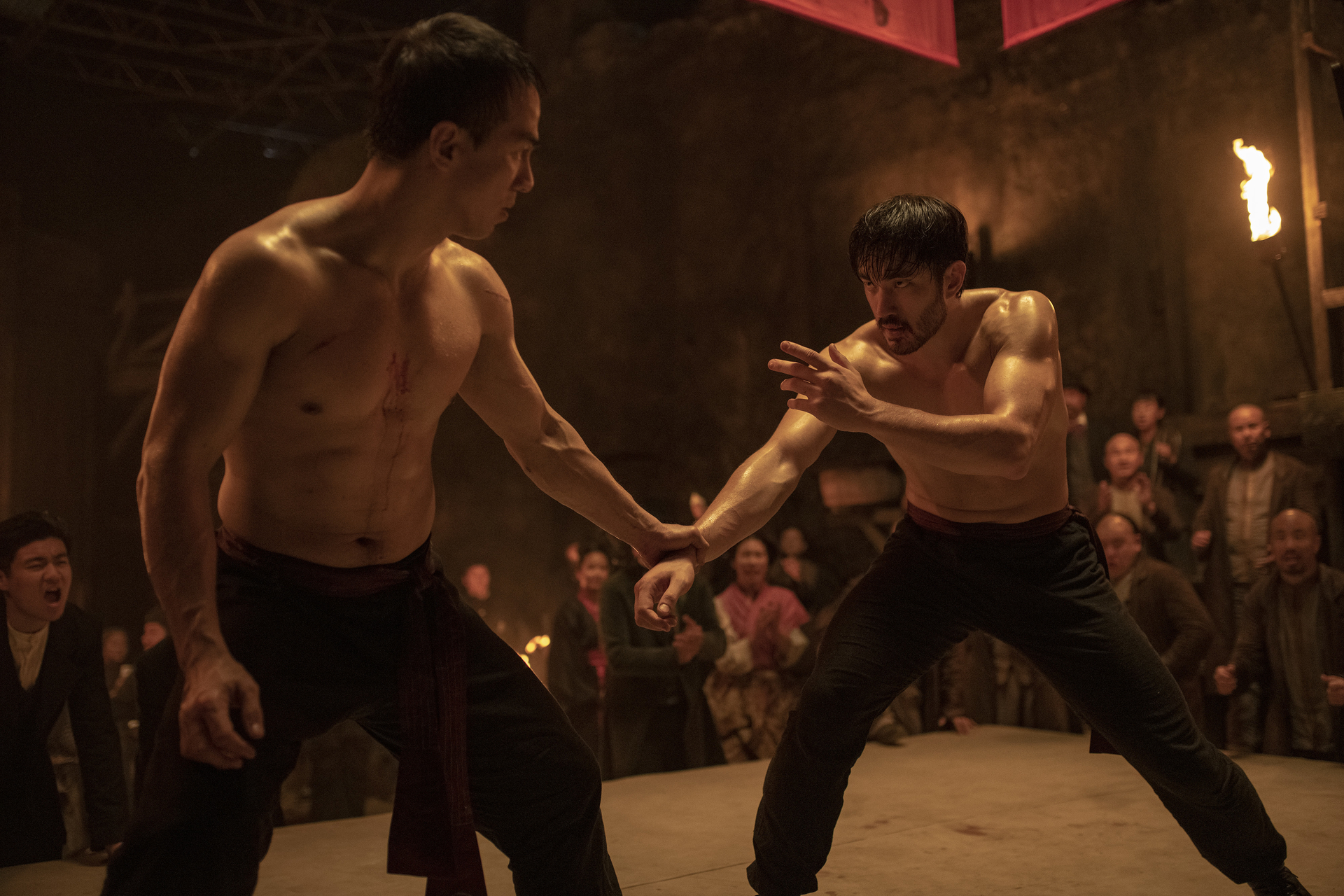Andrew Koji and Joe Taslim, both shirtless and drenched in sweat, grasp onto each other in fighting stances as excited onlookers watch in Warrior.