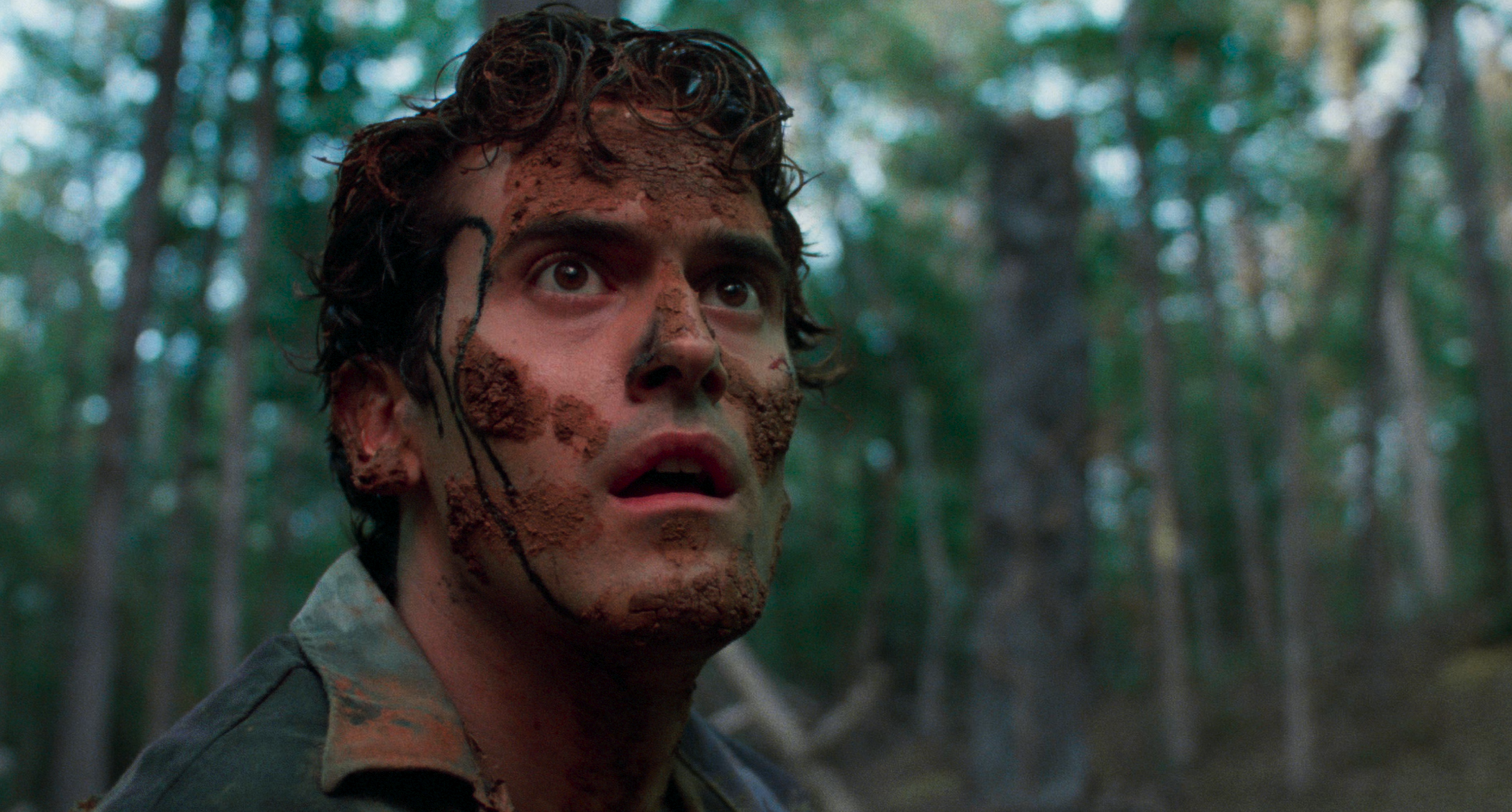 Bruce Campbell has dirt on his face and all in his hair while he looks up surprised in a forest in The Evil Dead.